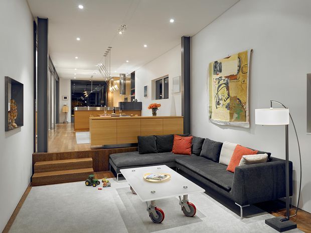 A contemporary open-plan living room flowing into a kitchen. The room features a large dark charcoal sectional sofa with orange-red cushions, a white coffee table on casters, a pale area rug, timber built-in cabinetry and bench seating, recessed ceiling lights, artwork, and a slim floor lamp. The kitchen is visible beyond a timber island and dark cabinetry, with a long, narrow layout and polished modern detailing.