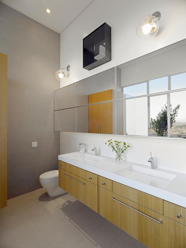 Bright contemporary bathroom with a long double vanity, white integrated basins and warm timber cabinetry. Large mirrored wall cabinets reflect the room and windows, while neutral large-format wall and floor tiles, modern wall lights, recessed ceiling lighting, and a toilet create a clean, refined interior. A wide window provides views toward a leafy urban landscape.