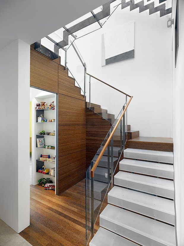 Bright modern stairway interior with a sculptural zigzag stair profile, white stair treads and risers, dark timber flooring and timber-clad joinery beneath the stairs. Clear glass balustrades with slim dark metal supports and a timber handrail surround the stair. A built-in open shelving niche with children's toys is visible beside the stair, while a large abstract artwork hangs on the white wall.