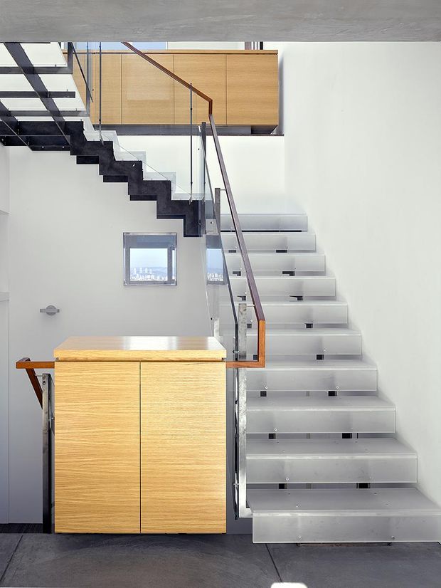 Interior architectural photograph centred on a modern stairway with floating grey stair treads, clear glass balustrades, and dark structural stringers. Warm wood-grain built-in cabinetry forms a landing or storage element beside the stairs, contrasting with white walls and a grey concrete ceiling and floor. The open, multi-level composition emphasizes the staircase and contemporary construction details.