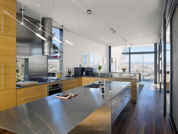 A bright, spacious contemporary kitchen with extensive warm timber cabinetry, dark grey metal benchtops, a large central island, stainless-steel appliances, and sculptural pendant lighting. Floor-to-ceiling glazing along the right side opens views toward a dense urban skyline, while dark timber flooring and white walls create a refined, high-contrast interior.