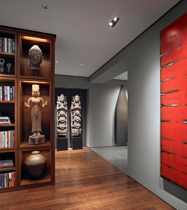 A refined interior hallway or gallery-like passage featuring dark timber built-in shelving filled with books and illuminated display niches for carved statues and pottery. The space has warm timber flooring, smooth grey walls and ceiling, recessed spot lighting, a large vivid red textured wall artwork or feature panel, and sculptural objects displayed along the corridor.
