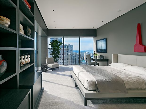 A bright, high-rise contemporary bedroom with a large upholstered platform bed, pale bedding, light carpet, and floor-to-ceiling glazing overlooking a dense cityscape and distant hills. Dark built-in open shelving occupies the left wall and displays decorative objects, while a compact desk and chair sit near the window. The room has a restrained neutral palette with a vivid red sculptural wall accent.