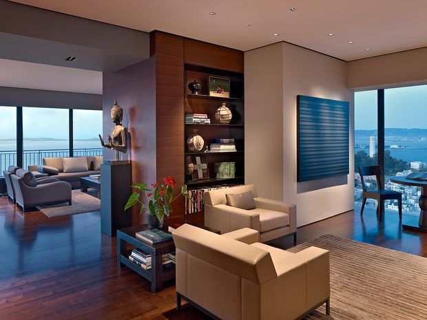 A sophisticated open-plan living room with warm wood flooring, built-in dark timber shelving, leather lounge chairs, and a central display wall. Floor-to-ceiling glazing opens views toward a coastal bay or waterfront setting, while a dining area is visible at right. The room has a refined contemporary feel with layered lighting and curated decorative objects.