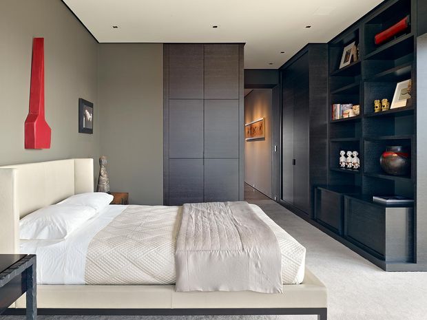 A refined contemporary bedroom with a low upholstered bed, white bedding, pale carpet, and a restrained grey, black, and white palette. Full-height dark timber or wood-look built-in cabinetry and open shelving line the right wall, while a large grey wardrobe panel occupies the rear wall. A bold red sculptural wall light provides the main accent. The room has a clean, calm, high-end appearance with recessed ceiling lighting and integrated joinery.