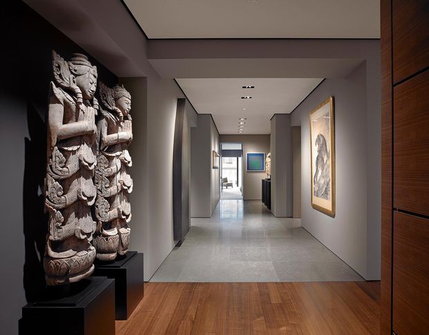 A refined interior hallway or entry passage with a warm timber floor, pale grey walls, recessed ceiling lighting, and dark timber built-in joinery on the right. A large carved sculptural artwork stands on a black plinth at the left, while framed artwork and a glimpse of a brighter room appear further down the corridor. The space has a gallery-like, contemporary character with strong contrast between the dark display wall, light corridor surfaces, and timber flooring.