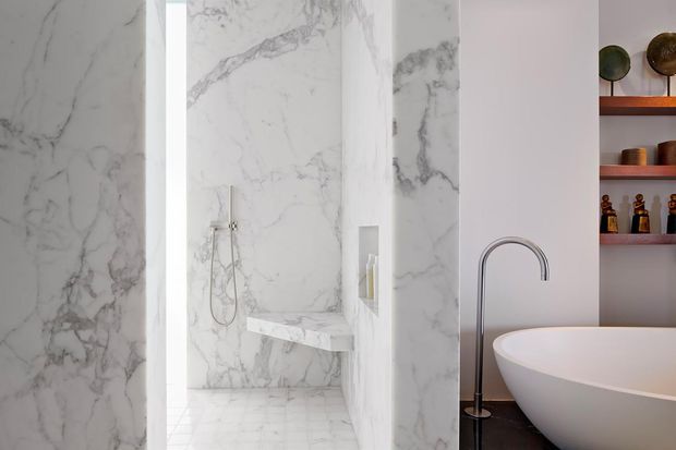A refined bathroom featuring a walk-in shower enclosed by dramatic white-and-grey veined marble surfaces, a marble shower bench and recessed niche, and a freestanding white oval bathtub with a tall chrome floor-mounted tap. Open timber shelving with decorative vessels is visible at the right.