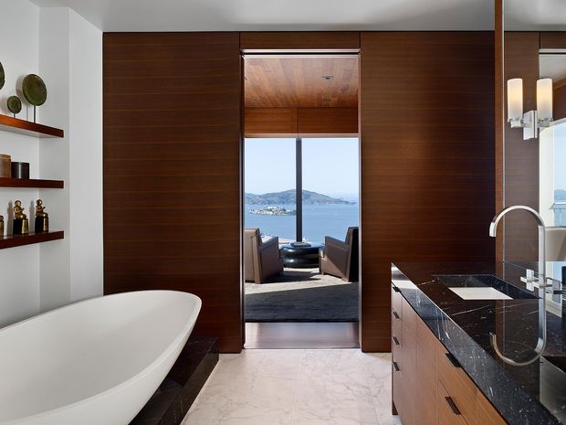 A luxurious contemporary bathroom with a freestanding white bathtub, dark timber wall panelling and concealed cabinetry, a polished dark stone vanity with integrated basin and tall curved tap, and pale marble flooring. Open double doors frame a view into a seating area and a blue waterfront landscape beyond. Shelving with decorative objects and wall-mounted cylindrical sconces add detail.