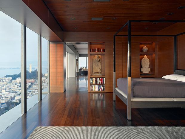 A sophisticated high-rise bedroom with a dark timber floor and matching timber ceiling and wall panelling. Floor-to-ceiling glazing provides expansive city views, while a four-poster bed with dark framing occupies the right side. Built-in timber shelving displays books and sculptural objects, and a corridor leads toward a small seating area in the background.