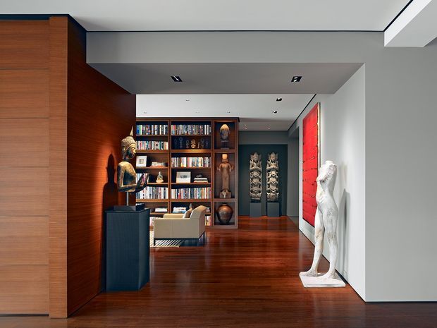 A refined modern interior hallway or gallery space with rich dark timber flooring and wall panelling, a large built-in bookcase displaying books and sculptures, recessed ceiling lights, and curated artwork and sculptural objects. The space opens toward a lounge or study area and uses strong contrasts between warm wood, charcoal walls, white surfaces, and a vivid red artwork panel.