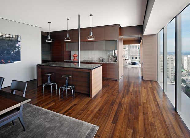 A refined open-plan kitchen and dining area in a high-rise apartment, featuring dark timber cabinetry, a large dark stone island with bar seating, pendant lights, stainless appliances, and rich timber flooring. Floor-to-ceiling glazing provides expansive urban views, while a dining table and chairs occupy the foreground.