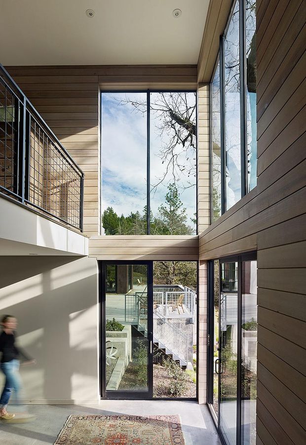 A bright, contemporary double-height interior entry or circulation space with extensive floor-to-ceiling glazing. Warm horizontal timber wall and ceiling lining contrasts with smooth light plaster walls, dark-framed glass doors, and a metal balustrade overlooking the lower level. Exterior decking, stairs, and landscaped surroundings are visible through the glazing. A patterned rug sits on the pale floor, while a person is partially visible at the edge of the frame.