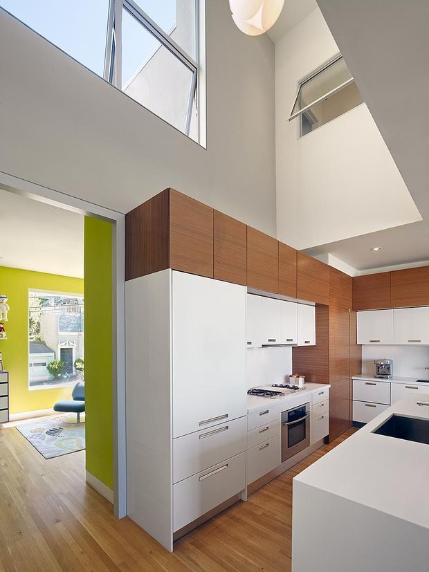 A bright, contemporary kitchen with tall white built-in cabinetry and warm wood-grain overhead and wall cabinetry. The space features a white island with a dark inset benchtop, integrated appliances, timber flooring, and a dramatic double-height ceiling with high-level glazing and an open void. A vivid lime-green adjoining room is visible through the wide opening.
