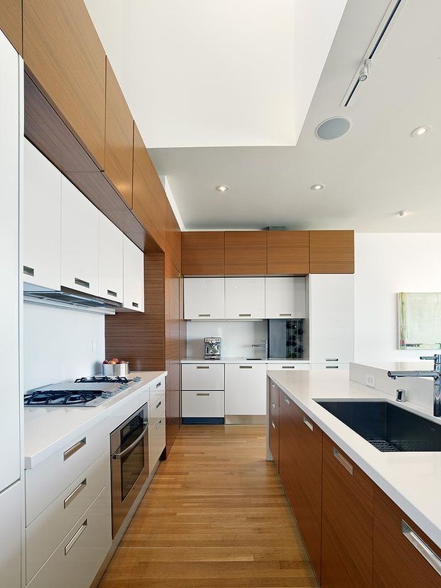 Bright contemporary kitchen with a galley-style layout and central island. White glossy cabinetry contrasts with warm horizontal timber wall and overhead cabinetry, while white stone-look benchtops, integrated appliances, a gas cooktop, and a deep dark sink create a clean, functional fit-out. Recessed downlights, a linear track light, and a partial bulkhead define the ceiling.