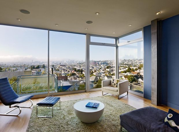 A contemporary living room with expansive floor-to-ceiling corner glazing overlooking a dense suburban cityscape. The room features a sculptural white round coffee table, pale textured rug, upholstered lounge chair and ottoman, dark lounge seating, timber flooring, recessed ceiling lights, and dark blue accent walls with a vertical stone-textured column. A balcony with a glass balustrade is visible beyond the glazing.