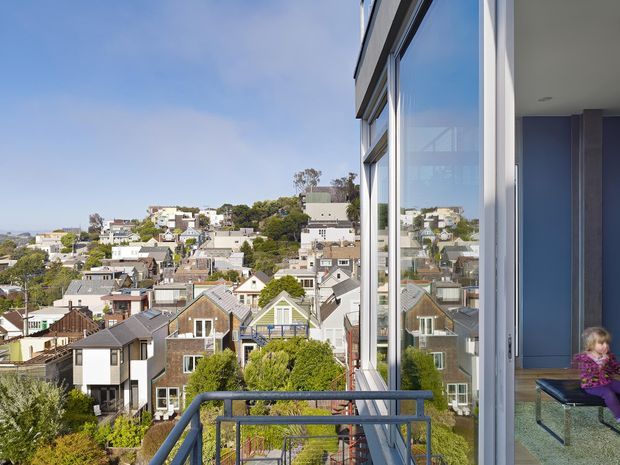 Exterior architectural view from a balcony or terrace, looking across a densely built hillside residential neighbourhood. The image prominently shows a modern building edge with expansive floor-to-ceiling glazing, light-coloured framing, and a metal balcony railing. An interior seating area and child are visible through the open glazing, but the primary subject is the exterior balcony and facade, with urban houses and a distant water or horizon view beyond.