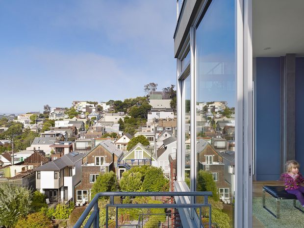 View from a contemporary home balcony across a dense, hilly suburban neighbourhood. The image is dominated by floor-to-ceiling glazing and a blue-grey metal balcony balustrade, with the interior edge of the home and a child visible at right. Numerous pitched-roof houses and mature trees fill the residential outlook under a clear sky.