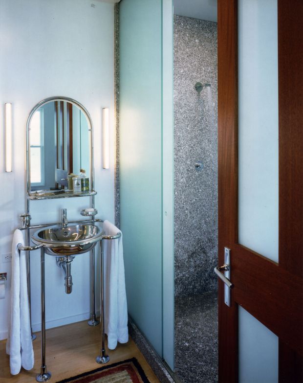 Compact bathroom with a chrome pedestal-style washbasin, exposed plumbing, arched mirrored medicine cabinet, wall-mounted vertical lights, pale blue walls, timber-look flooring, and a separate open shower lined in small grey stone mosaic. A dark timber-framed door with frosted glass is visible on the right.