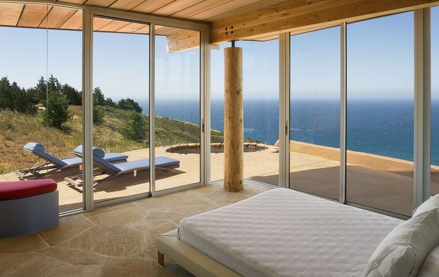 A bright coastal bedroom with a large bed in the foreground, framed by extensive floor-to-ceiling glazing and sliding glass doors. The room opens onto a sunlit terrace with loungers and expansive ocean views. Timber ceiling boards, exposed timber beams and a round timber support post contrast with the pale stone or tiled floor and neutral furnishings.