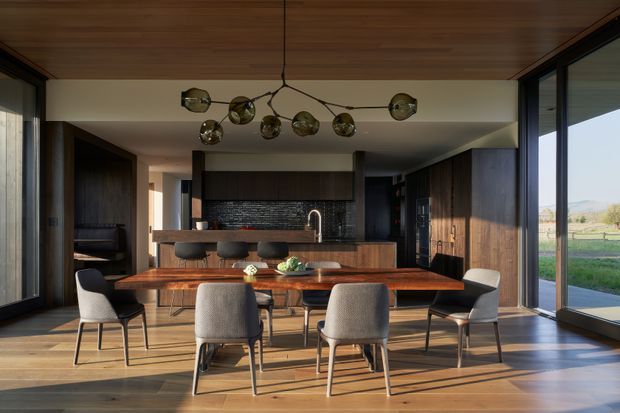 A refined open-plan dining and kitchen interior with a long timber dining table, upholstered dining chairs, a dark timber kitchen island, and black textured backsplash. Floor-to-ceiling glazing brings in views of the surrounding landscape, while a timber-lined ceiling and sculptural multi-globe pendant add warmth and visual interest.