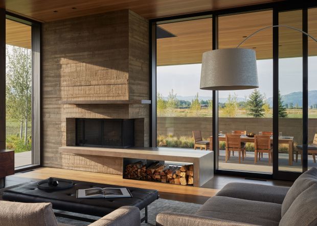 A warm contemporary living room centered on a substantial textured concrete fireplace with a long integrated hearth and open firewood storage. Full-height black-framed glazing connects the room to an outdoor dining terrace and landscaped mountain setting. Timber ceilings and flooring add warmth, while a large curved-arm pendant, low black ottoman, and sectional sofa furnish the space.