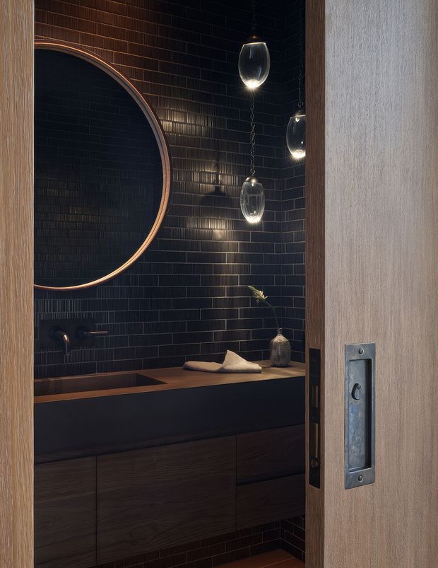 A dark, refined bathroom viewed through a partially open timber door. The room features a deep black tiled wall, a large round mirror with a warm metallic rim, a dark rectangular vanity with integrated basin and timber cabinetry, wall-mounted tapware, and several suspended glass pendant lights. The palette is moody and warm, with bronze and dark wood details.