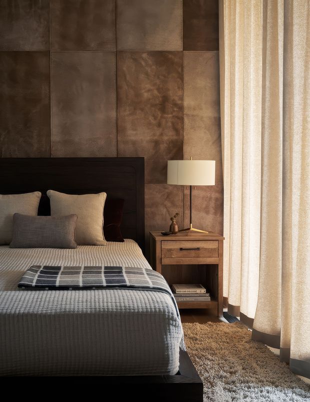 A warm, moody bedroom interior featuring a dark timber bed with layered neutral bedding, a large upholstered or leather-look brown wall panel feature behind the bed, and a timber bedside table with a table lamp. Full-height sheer curtains filter daylight along the right side, while a deep textured rug covers the floor.