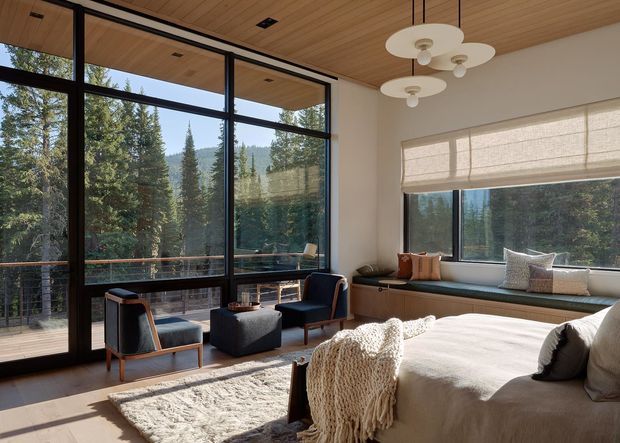 A serene, light-filled bedroom with expansive black-framed floor-to-ceiling glazing overlooking a forested mountainous landscape. The room features a built-in window seat, timber-lined ceiling, neutral upholstered bed, textured throw, sculptural pendant lighting, and a pair of mid-century-inspired lounge chairs arranged on a pale rug.