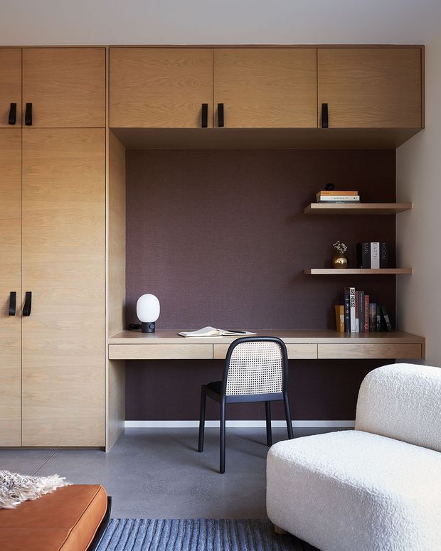 A compact, carefully designed home office with a built-in timber desk, extensive matching storage cabinetry, open display shelves, and a dark mauve textured feature wall. A black-framed cane-back chair sits at the desk, with a sculptural white table lamp, books, and a partially visible boucle armchair and leather ottoman adding warmth and texture. The room has a refined, contemporary character with a muted, warm palette.