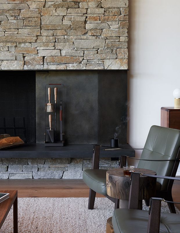 A refined living room centered on a substantial stacked-stone fireplace with a dark metal firebox surround and hearth shelf. Mid-century-inspired leather lounge chairs, a rustic round wood side table, dark timber furniture, a textured neutral rug, and a small timber cabinet create a warm, moody seating area. The composition emphasizes natural stone, dark metal, timber, and muted olive-green upholstery.