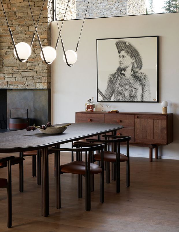 A refined dining room with a long dark dining table and matching dark wood and leather chairs. A warm timber sideboard sits against a pale plaster wall beneath a large monochrome portrait, while a stacked-stone feature wall and dark fireplace add texture. Sculptural globe pendant lights hang above the table, and the room has timber flooring with a warm, moody contemporary character.