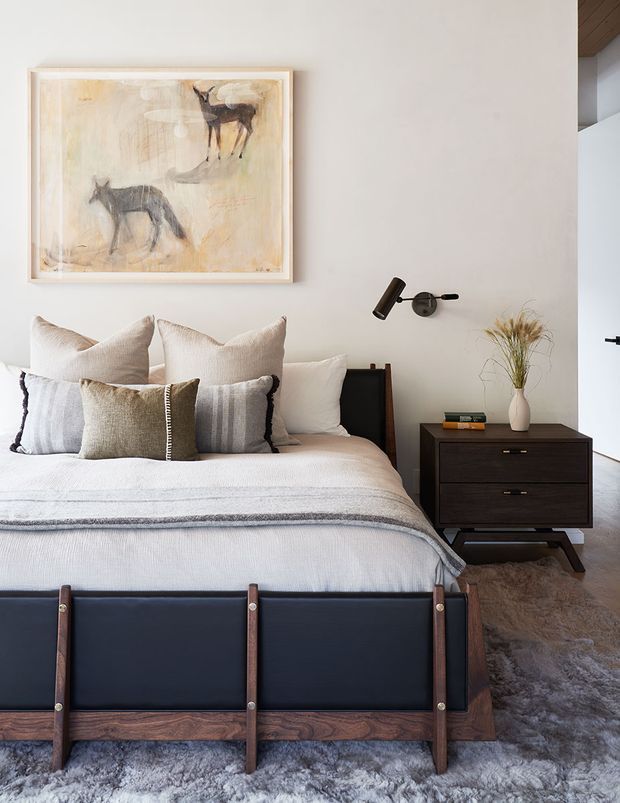 A warmly styled bedroom with a large upholstered bed, layered neutral and striped bedding, and a dark timber-and-black leather bed frame. A matching dark timber bedside cabinet sits beneath a black wall-mounted reading light, with a framed animal artwork above the bed. A textured grey rug covers the floor, while a white wall and partial timber ceiling add understated architectural warmth.