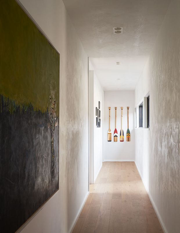 A narrow, bright residential hallway with textured white plaster walls, pale timber floorboards, recessed ceiling lights, and a small window at the far end. Decorative paddles are mounted on the end wall, while a large abstract artwork occupies the left wall. The space has a clean, understated contemporary feel with natural timber accents.