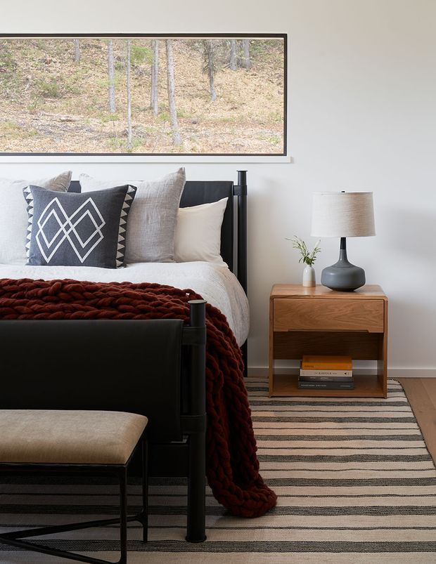 A refined bedroom interior with a black four-poster bed, layered neutral bedding, a chunky rust-red knitted throw, and patterned geometric cushions. A warm wood bedside table holds a dark ceramic lamp and small vase, while a striped neutral rug spans the floor. A large framed artwork or window-like forest image hangs above the bed, contributing an earthy, calm atmosphere.