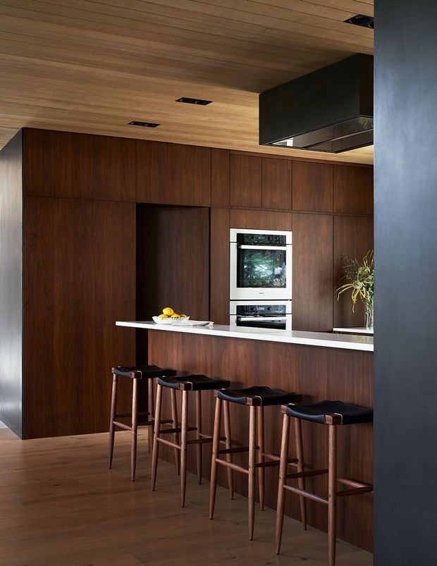 A refined contemporary kitchen with extensive dark timber cabinetry and matching timber ceiling and flooring. A white stone island benchtop provides contrast, with four bar stools along the island. Integrated double ovens, a concealed doorway, recessed ceiling lights, and a dark rangehood enclosure complete the streamlined built-in design.