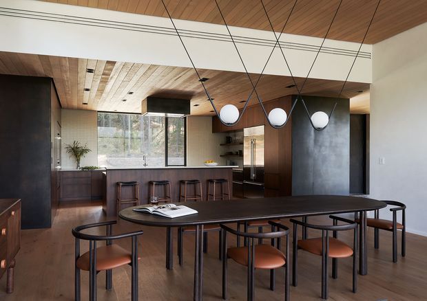Open-plan dining and kitchen interior with a long dark timber dining table and coordinated chairs in the foreground. The kitchen includes a dark timber island with bar stools, integrated cabinetry, a large window, and a dark stone or concrete feature wall. Warm timber lining continues across the ceiling, complemented by white structural beams and sculptural globe pendant lights suspended on black cords.