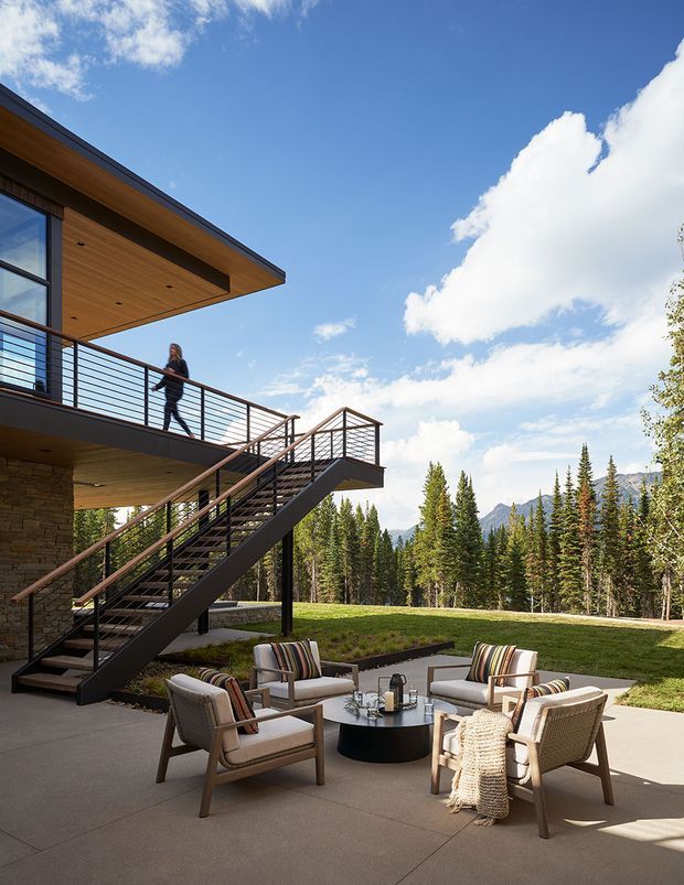 Contemporary multi-level house exterior with a broad timber-lined roof overhang, stone-clad lower wall, and dark metal-framed glazing. An elevated balcony and open steel stair provide access to a paved outdoor seating area with lounge chairs and a fire-pit-style table. The property is set among coniferous woodland with mountain views under a bright blue sky.