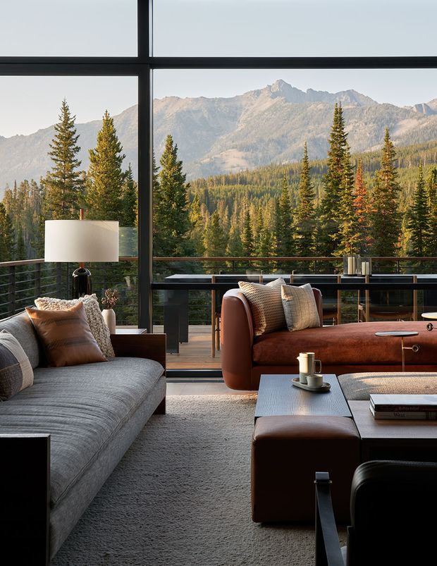 A refined living room with floor-to-ceiling black-framed glazing opening onto a balcony and expansive mountain and forest views. The room features a large grey upholstered sofa, rust-brown leather sofa, textured cushions, a dark coffee table, leather ottoman, sculptural lamp, and a restrained palette of charcoal, grey, brown, and warm timber tones. The glazing and balcony create a strong connection to the alpine landscape.