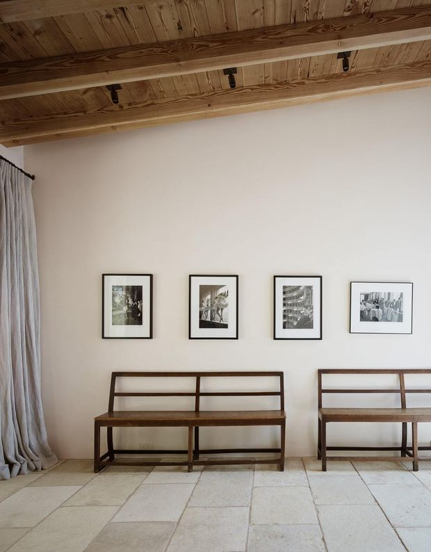 A calm, minimalist interior with a high exposed timber ceiling, white plaster walls, pale large-format stone or concrete-look flooring, and two dark timber open-back benches positioned beneath a row of framed black-and-white photographs. Full-height curtains hang at the left edge of the room. The space has a restrained, gallery-like character with warm natural timber detailing.