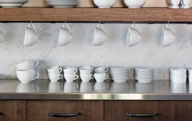 Close-up of a built-in serving cupboard displaying neatly arranged white cups, saucers, bowls and plates. The unit features dark timber cabinetry and shelving, a polished metal counter edge, and a pale grey-white marble or stone backsplash. White cups hang from hooks beneath the upper timber shelf.