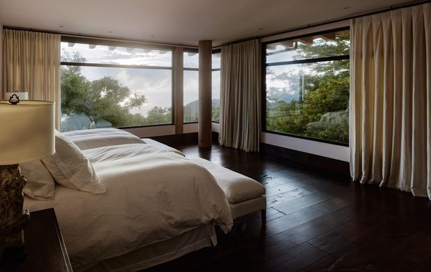 A refined bedroom with a large upholstered bed, bedside lamp, dark timber flooring, and full-height corner glazing framed by flowing cream curtains. The windows provide leafy outdoor views and abundant natural light, while a central timber-clad column and dark trim add architectural definition.