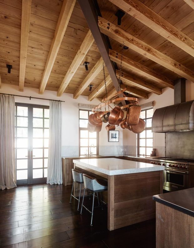 A warm, rustic kitchen with a large central island, dark timber cabinetry, stone benchtop, stainless-steel rangehood and cooker, and stools tucked beneath the island overhang. The room features exposed timber beams and rafters across a pitched ceiling, timber flooring, tall multi-pane glazed doors and windows with curtains, and a prominent hanging rack of copper cookware.