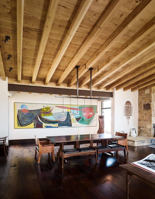 A warm, rustic dining room with a long dark wood dining table and matching wooden chairs beneath an exposed timber-beam and timber-plank ceiling. A large colourful abstract artwork spans the white wall, while a substantial natural stone fireplace or masonry feature occupies the right side. Dark timber flooring, black linear lighting, and expansive glazing contribute to the inviting, architecturally detailed interior.