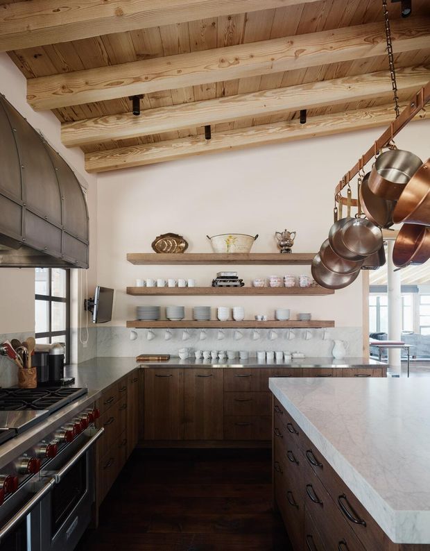 A warm, rustic kitchen with dark wood cabinetry, a large pale stone island, open timber shelving, and a pale tiled backsplash. Copper and stainless-steel cookware hangs prominently from an overhead rack. The room features a timber-lined ceiling with exposed beams, dark timber flooring, a large range cooker, and generous glazing at the sides.
