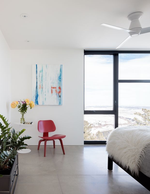 Bright, minimalist bedroom with white walls and ceiling, a polished light-grey concrete or large-format tiled floor, and full-height black-framed glazing overlooking a coastal landscape. The room includes a low black bed with a fluffy white throw, a sculptural red accent chair, indoor plants, a small side table with flowers, abstract artwork, recessed lighting, and a contemporary ceiling fan.