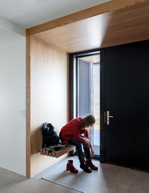 A contemporary residential entryway with a built-in light timber-lined alcove and bench beside a large matte-black entry door. A person is seated putting on boots, with coats and bags on the bench. The space has pale walls, a concrete-look floor, a neutral textured rug, black-framed glazing, and strong natural light entering through the open doorway.