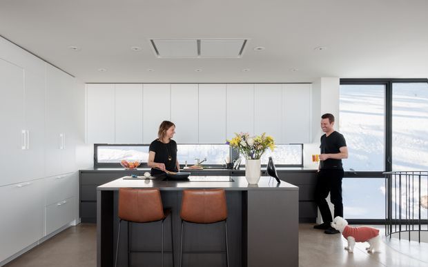 Bright contemporary kitchen with a large dark island, two leather bar stools, extensive white full-height cabinetry, integrated appliances, and a broad window overlooking a snowy landscape. Two people and a small dog are present, with a floral arrangement on the island. The space has a clean, minimalist fit-out and a stair railing visible at right.