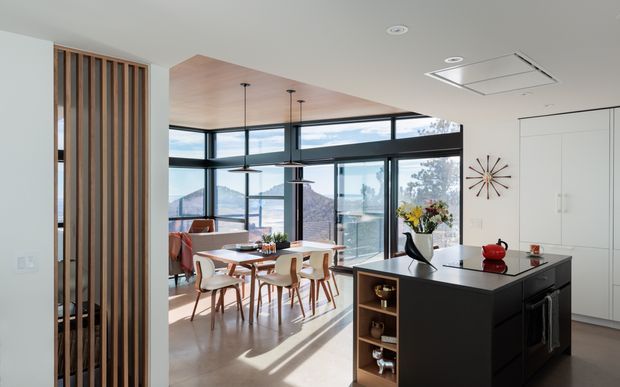 A bright, open-plan contemporary kitchen and dining area with a black island, integrated white cabinetry, timber dining table and chairs, and a glazed corner overlooking a distant landscape. A timber slatted screen separates the space from the entry, while pendant lights hang beneath a warm timber ceiling feature.
