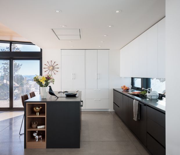 A bright contemporary kitchen with a dark island featuring open timber display shelving, integrated dark cabinetry, white upper cabinets, and a long black benchtop. Floor-to-ceiling glazing and a glass door bring in abundant natural light and views beyond. Recessed ceiling lights, a linear skylight or ceiling vent, pendant-free clean lines, and restrained decor create a refined modern interior.