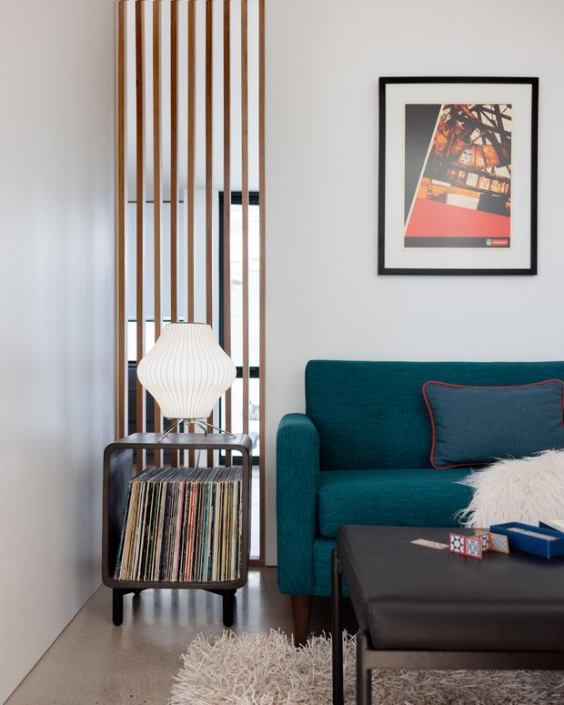 A stylish living room corner with a teal upholstered sofa, black leather coffee table, shag rugs, and a vintage record-player cabinet filled with vinyl records. A sculptural white pendant lamp sits above the cabinet, while vertical timber battens screen a glazed opening behind it. The pale walls and concrete-look floor create a clean modern backdrop, complemented by framed graphic artwork and dark furniture accents.